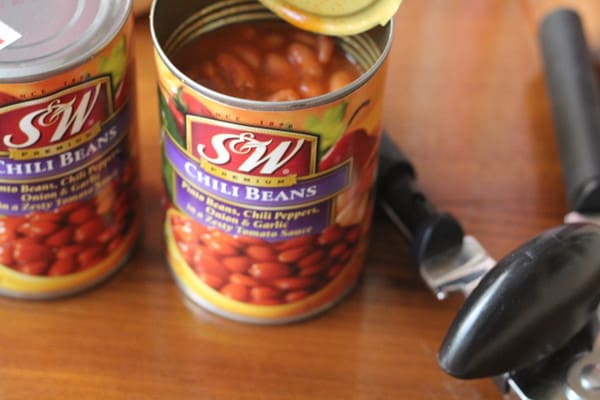 two cans of chili beans on a wooden table next to a manual can opener